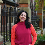 A photograph of a woman in a brightly coloured jumper. The woman is smiling at the camera.