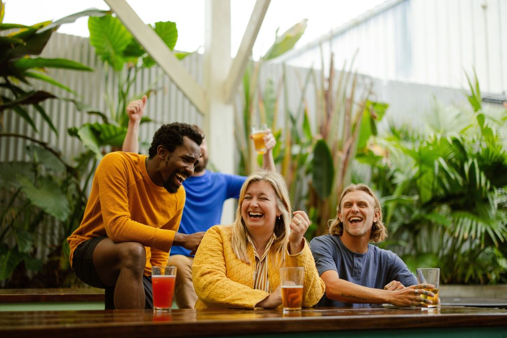 Group of friends laughing together at outdoor bar, wearing yellow and blue casual clothes, surrounded by tropical plants