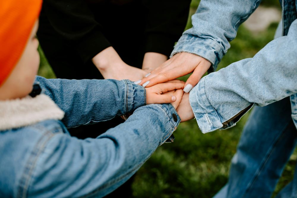 Close-up of several hands stacked together, all wearing denim sleeves, against a grassy background