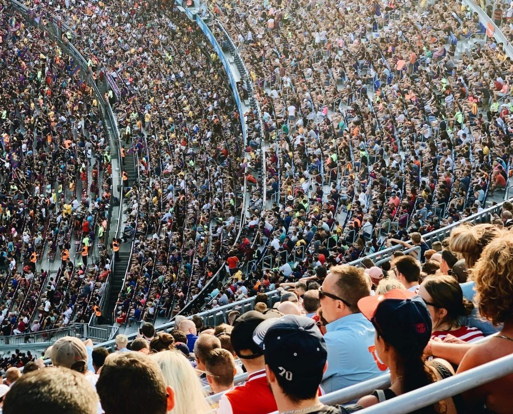 Packed stadium seating filled with thousands of spectators, viewed from above showing curved rows of seats and crowd in casual attire