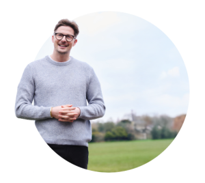 Smiling man wearing glasses and grey sweater standing in front of grassy field with buildings in distance