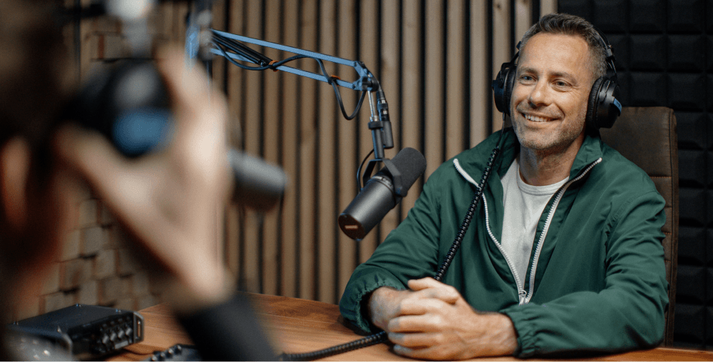 Person in green jacket smiling while recording a podcast, wearing headphones with a professional microphone setup on boom arm