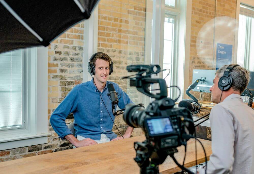 Two people recording a video in a studio with exposed brick walls, wearing headphones at a wooden table, with professional camera equipment set up to film