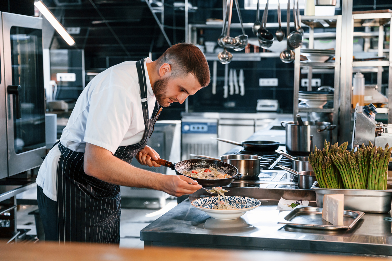 Chef in striped apron plating a dish in professional kitchen with fresh asparagus nearby