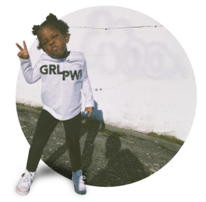 Young child in white 'GRL PWR' shirt and black pants making peace sign and smiling against white wall