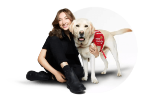 Person in black clothing sitting next to a yellow Labrador wearing a red service dog vest, both looking happy