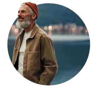 Smiling middle-aged man with gray beard wearing orange beanie, white t-shirt and brown work jacket, photographed against blurred waterfront with evening lights