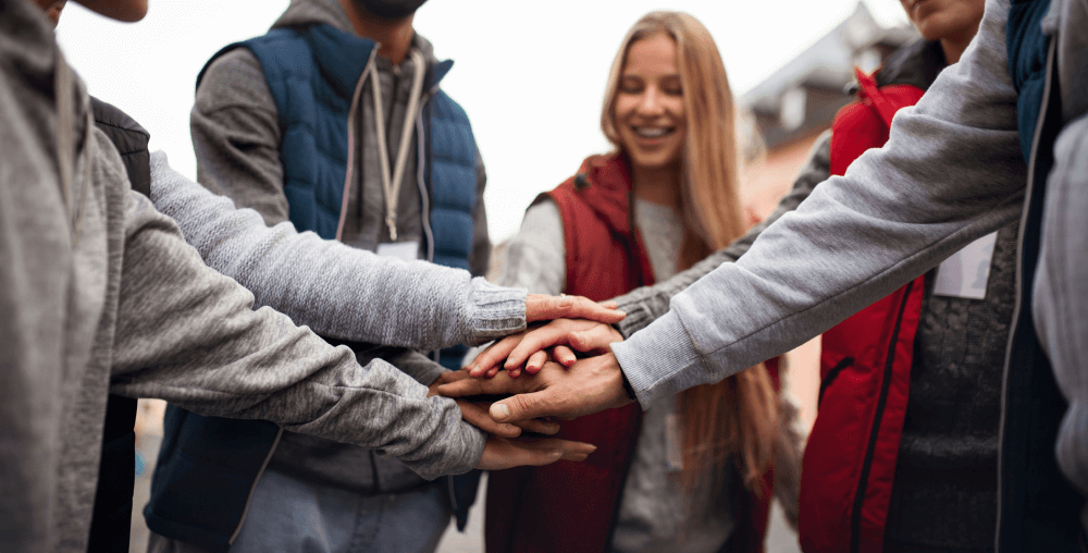 Group of young people in casual winter wear putting hands together in a team huddle, with a smiling woman in a red vest visible in the background