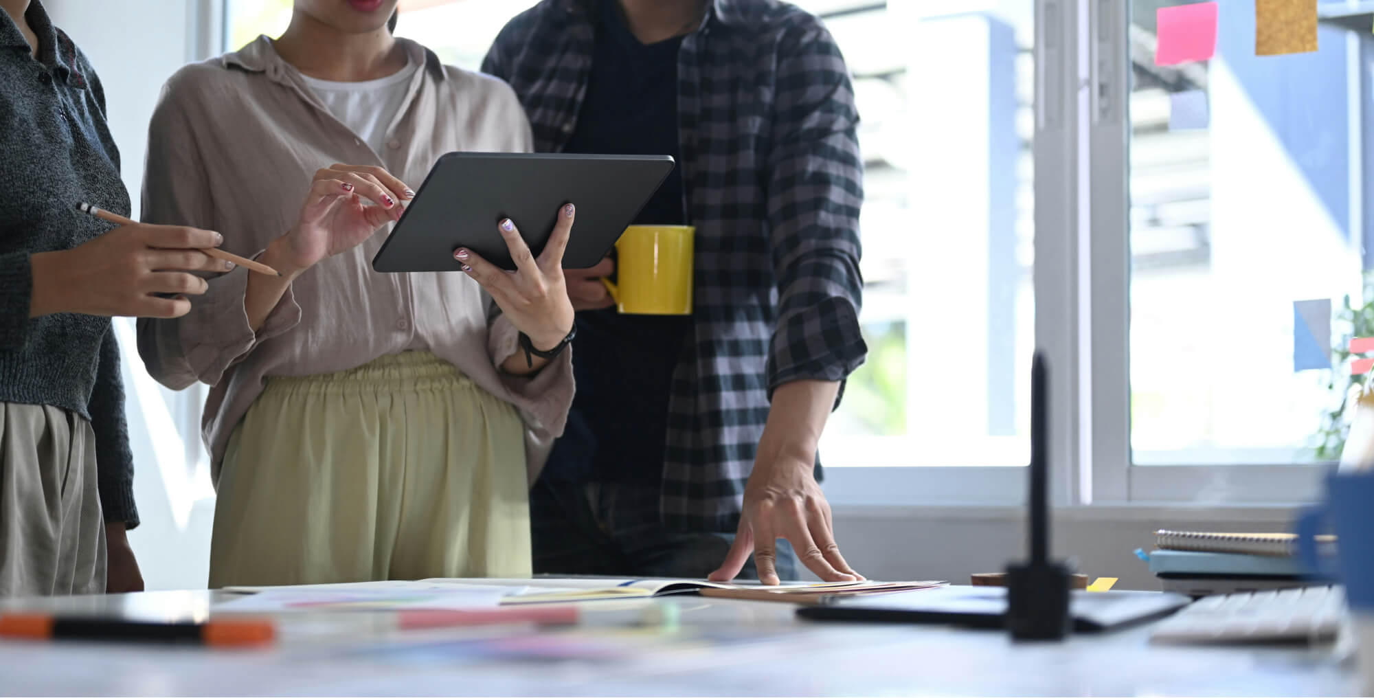 People collaborating around a desk, looking at a tablet and documents, with one person holding a yellow coffee mug