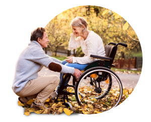 Senior couple sharing tender moment in autumn park, man kneeling beside woman in wheelchair, both wearing knit sweaters with fallen leaves around them