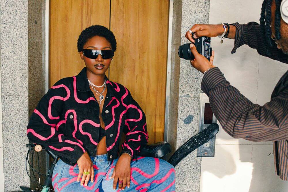 ehind-the-scenes photo of a fashion shoot - model wearing black and pink patterned jacket with matching jeans and sunglasses poses while photographer takes picture