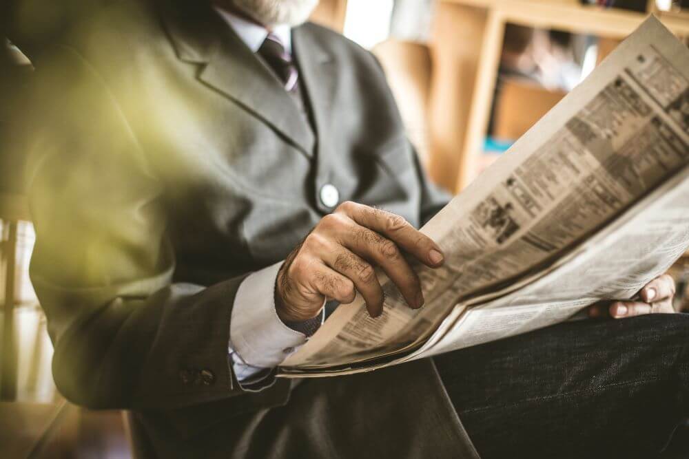 Close-up of hands holding a newspaper, person wearing a dark business suit with white shirt and tie