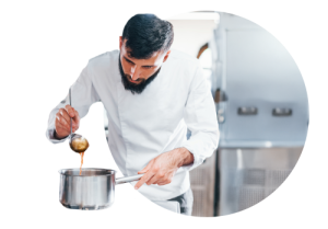 Chef in white uniform carefully pouring liquid from a ladle into a metal pot in a professional kitchen