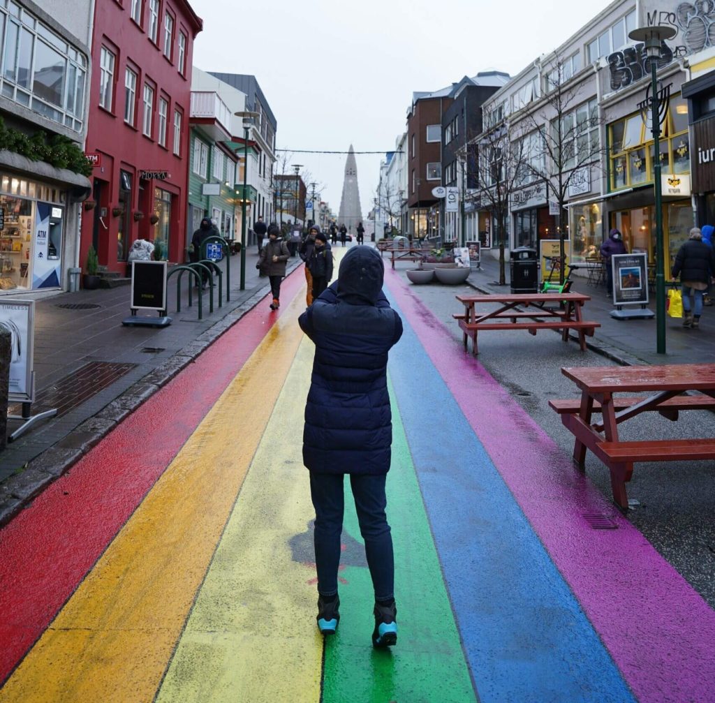 Person in dark winter coat walking on rainbow-colored painted street with shops and a church spire visible in the distance