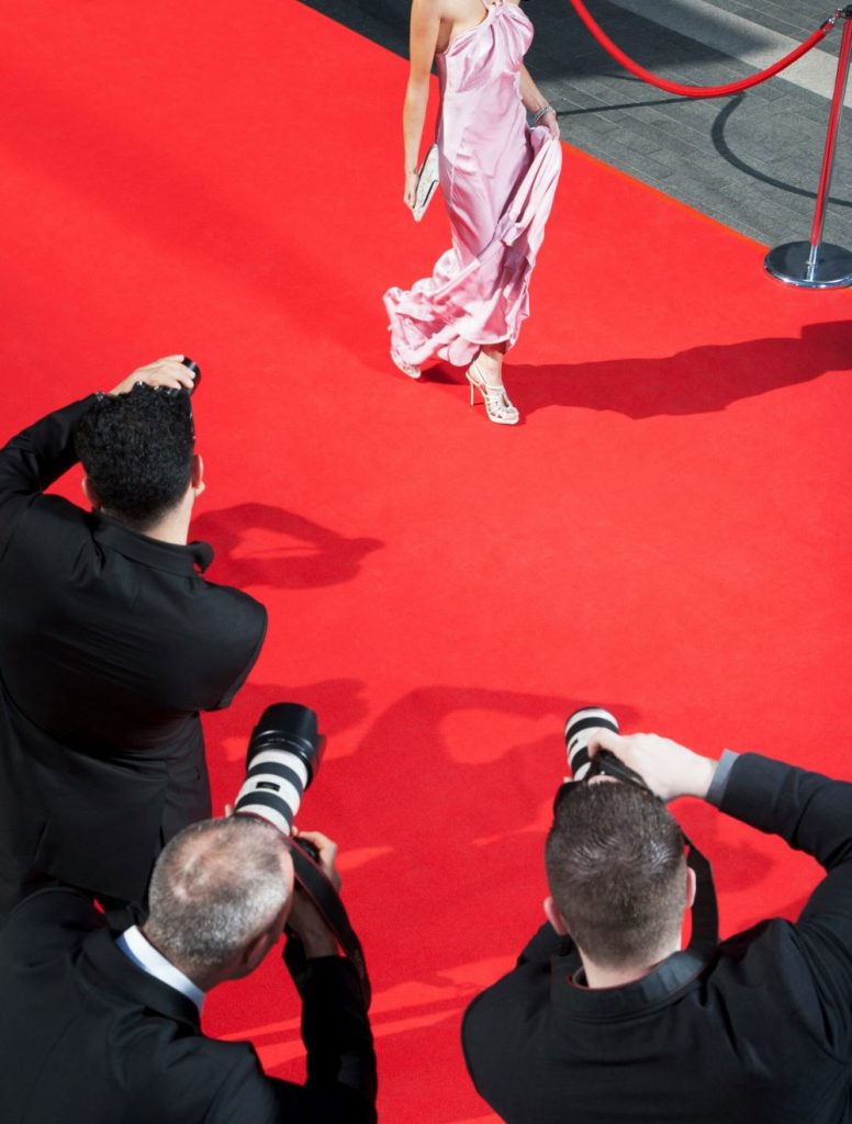 Photographers shooting a person in pink gown walking on red carpet, viewed from above showing three photographers with professional cameras in black attire