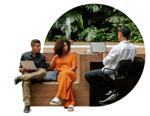 Three people having casual meeting outdoors, sitting on wooden bench with laptops, woman in orange dress between two men, tropical plants in background