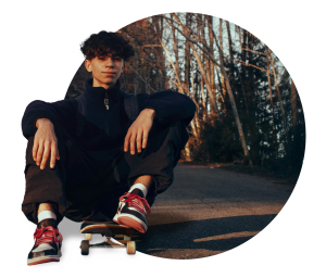 Young person with curly hair sitting on skateboard at sunset, wearing black athletic wear and red trainers