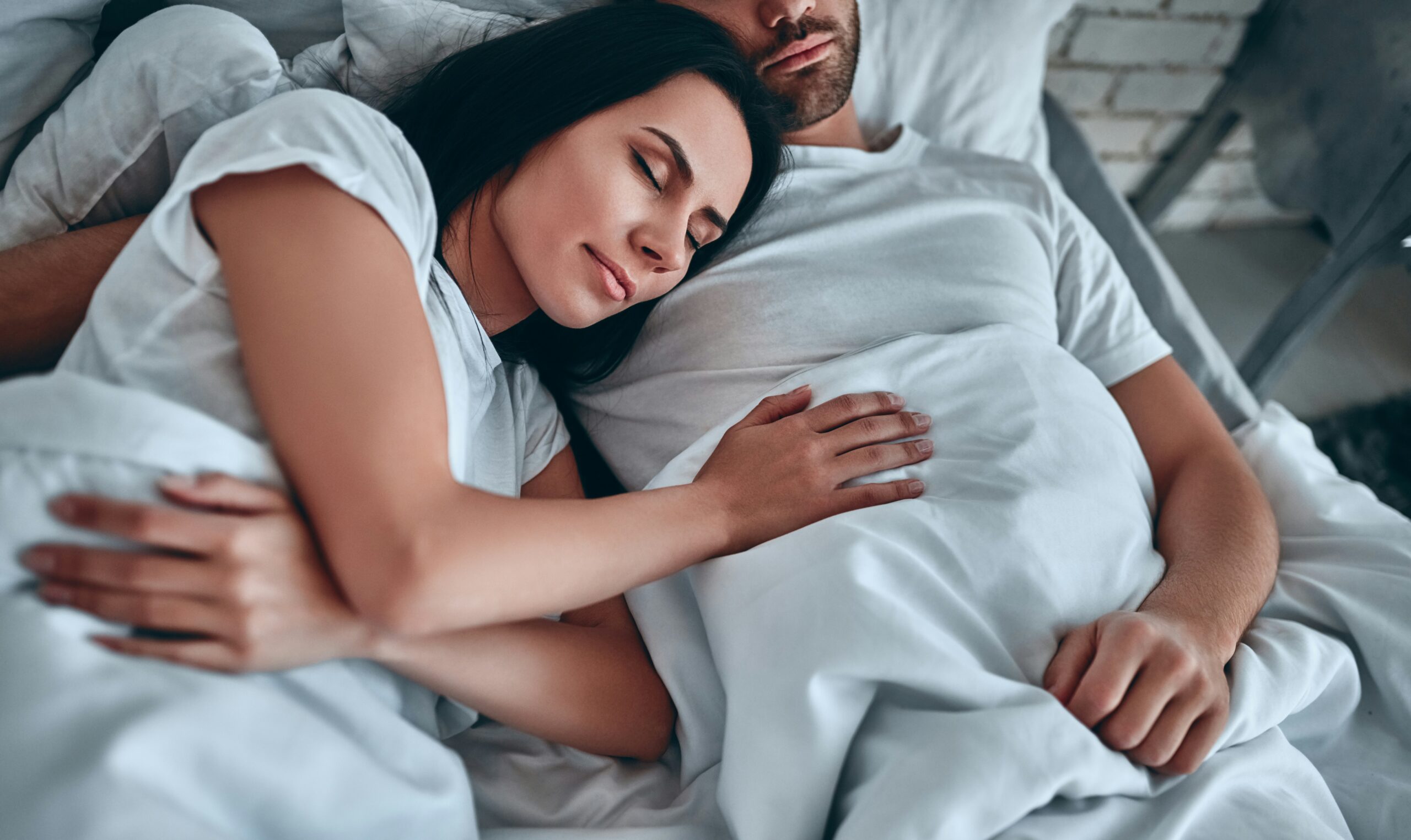 Couple sleeping peacefully in white bedding, woman resting head on partner's chest