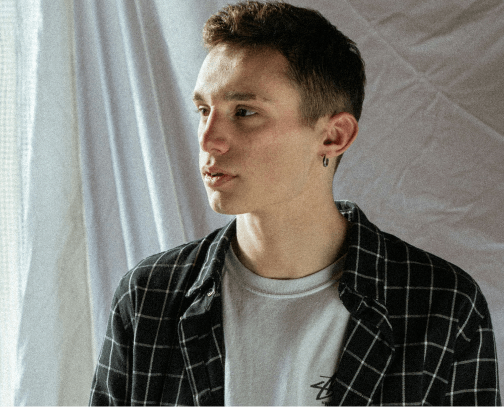 Side profile portrait of young person wearing plaid overshirt and white t-shirt against white curtain background