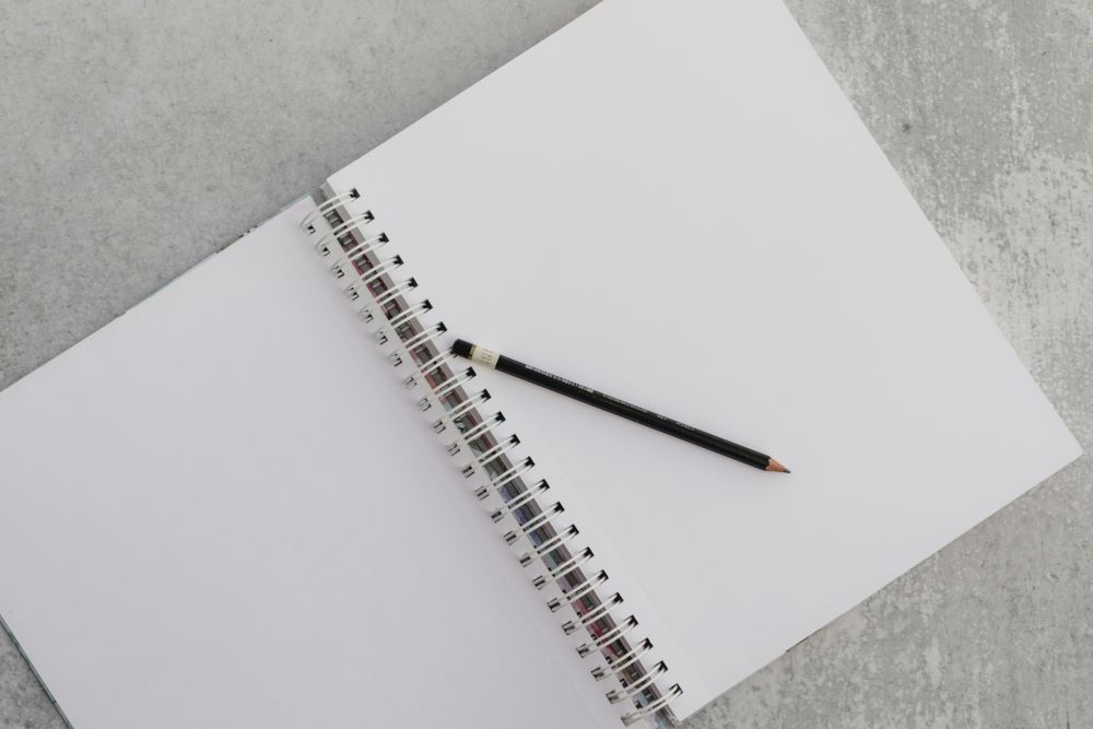 Black pencil lying diagonally across an open blank spiral notebook on a grey surface